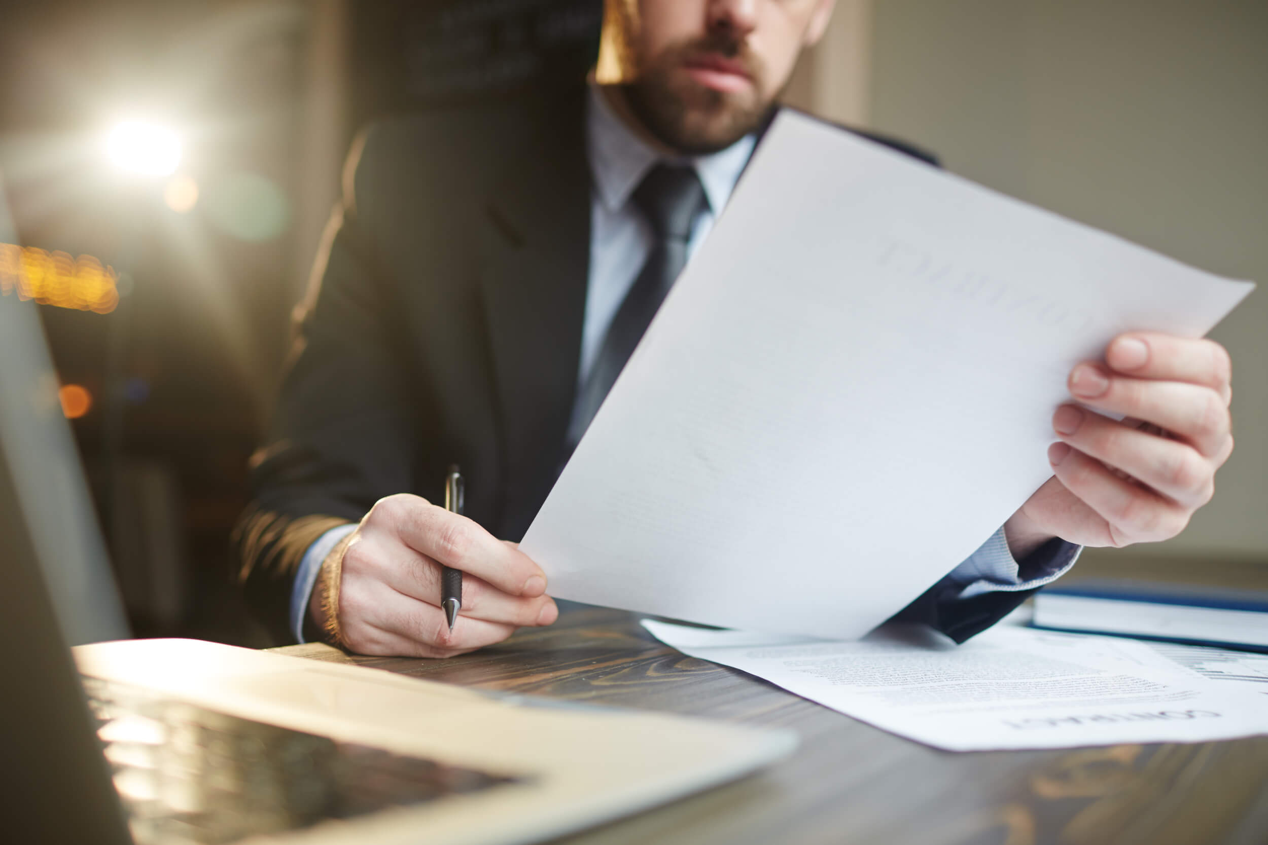 Portrait of modern bearded businessman holding papers in hands, reading and analyzing contract documentation at desk with laptop krypto-advokat-bratislava-slovakia