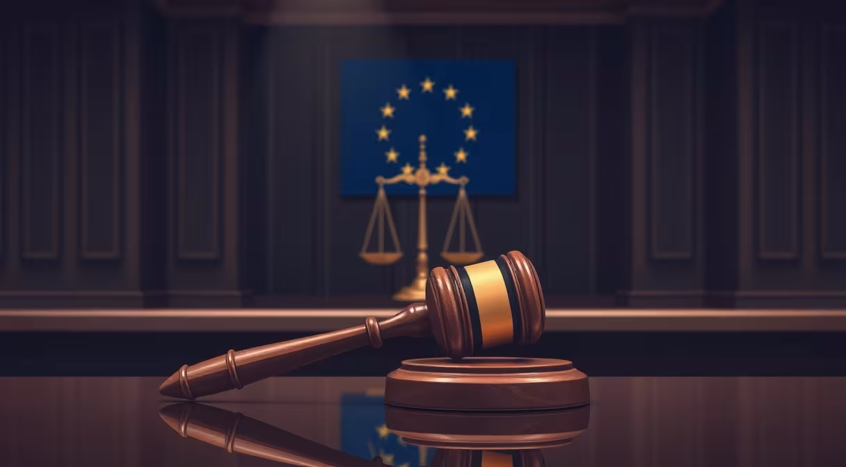 Sudcovské kladivo na stole s váhami spravodlivosti a vlajkou EÚ v pozadí, symbolizujúce právomoci a sankcie.